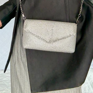 Glittrig silver kuvertväska med kedja - Säljer en glittrig kuvertväska/clutch i silver med kedjerem. Väskan är täckt av små glittrande stenar och har en stilren, rektangulär form med lock. Perfekt för dig som vill sticka ut och addera extra bling till din outfit. Ny