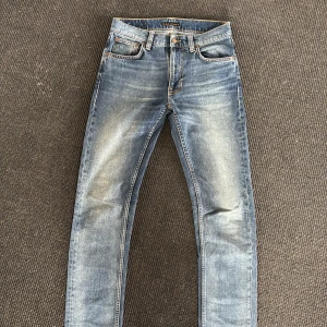 Blå skinny jeans från Nudie Jeans - Snygga blå skinny jeans från Nudie Jeans med klassisk femficksdesign och slitna detaljer på låren och knäna. Jeansen har ljus tvätt och kontrastsömmar, samt Nudie-logga på bakfickan. Perfekta för en avslappnad och trendig look.