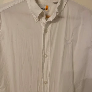 Vit skjorta från Timberland M - Vit skjorta från Timberland i relaxed fit med klassisk krage och knappar framtill. Skjortan har broderad logga på bröstet och är tillverkad i mjuk bomull. Perfekt för en clean och stilren look.