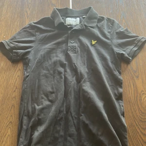Svart pikétröja från Lyle & Scott - Svart pikétröja från Lyle & Scott med klassisk krage och två knappar framtill. T-shirten har korta ärmar och är gjord i mjuk bomull. På bröstet sitter den ikoniska gula fågel-loggan som ger en snygg touch. Perfekt för en chill och stilren look.