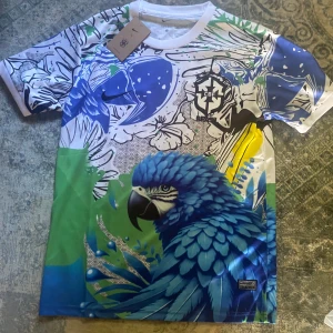 Färgstark CBF t-shirt med papegojtryck - Unik t-shirt från CBF med ett stort blått papegojmotiv, tropiska blommor och blad i blått, grönt, vitt och svart. Tryckt logga på bröstet och coola grafiska detaljer. Skön polyesterkvalitet och korta ärmar, perfekt för dig som gillar att sticka ut.