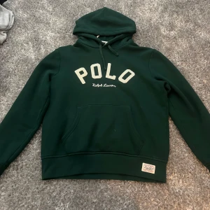 Mörkgrön hoodie från Polo Ralph Lauren - Mörkgrön hoodie från Polo Ralph Lauren med stor vit POLO-text på bröstet och klassisk känguruficka. Tröjan har huva med snörning. Perfekt för en chill och stilren look. Nypris-2400kr/ mitt pris-1400