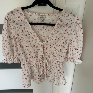 Blommig blus med knyt från AWARE, Vero Moda - Supersöt blommig blus från AWARE i vit med små rosa och röda blommor. Blusen har v-ringning, knyt framtill, puffiga korta ärmar och smock i midjan för en feminin siluett. Perfekt för vår och sommar när du vill ha en romantisk vibe.