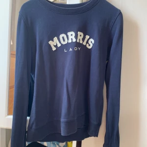 Mörkblå sweatshirt från Morris Lady - Mörkblå sweatshirt från Morris Lady med vit broderad logga framtill. Klassisk rund halsringning och långa ärmar. Skön och stilren tröja i bomull, perfekt för en avslappnad look.