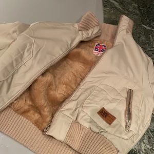 Beige bomberjacka från Billybay - Snygg beige bomberjacka från Billybay med mjukt fuskpälsfoder i brunt. Jackan har ribbade muddar, dragkedja framtill och en ärmficka med dragkedja. Perfekt för kyliga dagar och ger en cool, avslappnad look. 