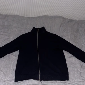 Mörkblå stickad kofta från Selected Homme - Mörkblå stickad kofta från Selected Homme med hel dragkedja framtill och hög krage. Klassisk och stilren modell i mjuk bomullsblandning, perfekt för lager-på-lager under kyliga dagar. Långärmad och ribbade muddar vid ärmslut och nederkant.