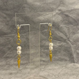 Guldiga örhängen med pärlor och spikes - Snygga örhängen med guldfärgade detaljer, två vita pärlor på rad och en liten guldig spike längst ner. Hängande modell som ger en cool och elegant vibe. Perfekt för dig som vill sticka ut med accessoarer som kombinerar klassiskt och edgy.