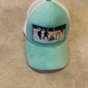 Turkos och vit keps med hockeymotiv - Säljer en keps i turkos och vitt med mesh på sidorna och baksidan. Framsidan har ett stort tygmärke med ett hockeymotiv. Kepsen har böjd skärm och är tillverkad i syntetmaterial och mesh för en luftig känsla.
