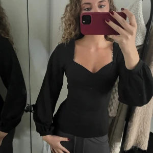 Svart topp med v-ringning  - Säljer en svart långärmad topp. Toppen har v-ringad hals, markerad midja och långa puffiga ärmar. Knyte vid ryggen