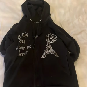 Svart hoodie med strass från Blinglucid - Svart hoodie från Blinglucid i storlek XS med dragkedja och huva. Framsidan har strassdetaljer med texten 'paris au clair de lune' och en glittrig Eiffeltorn-design. Perfekt för dig som gillar statement-plagg och snygga detaljer.