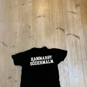 Svart Hammarby t-shirt med tryck - Svart t-shirt med vit text 'HAMMARBY SÖDERMALM' på ryggen och Hammarby-emblem i grönt och gult på bröstet. Klassisk rund hals och korta ärmar, perfekt för dig som vill visa ditt stöd för Hammarby. Skön bomullskvalitet och stilren design.