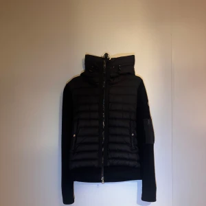 Moncler Cardigan - Säljer en svart pufferjacka från Moncler med stickade ärmar och huva. Jackan har dragkedja framtill, ficka med dragkedja på ärmen och Moncler-logga. Perfekt för sommaren och våren och hösten. Tillkommer med Moncler påse  samt den tags. NFC-Scan funkar (äkta)!          Mvh Tamana🤍🤗 var inte rädd för att fråga om fler bilder. OBS KONTAKTA INNAN KÖP