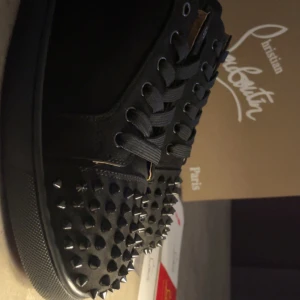 Christian Louboutin svarta sneakers med nitar - Svarta sneakers från Christian Louboutin med coola svarta nitar över tån och snörning. Skorna har en platt sula och är tillverkade i skinn och textil. Medföljer originalpåse, extra nitar och äkthetskort. Perfekt för dig som vill sticka ut med en edgy stil.
