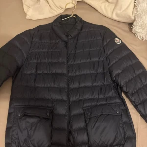 Svart dunjacka från Moncler - Säljer en svart dunjacka från Moncler med klassisk quiltad design och diskret logga på ärmen. Jackan har hög krage, dragkedja framtill och två stora fickor med lock och knappar. Perfekt för kalla dagar och riktigt snygg till streetwear.