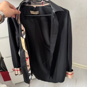 Svart Burberry skjorta med rutiga detaljer - Svart skjorta från Burberry i bomull med klassiska beige och röda rutiga detaljer på insidan av kragen, knappslån och ärmsluten. Skjortan har knappar framtill och lång ärm, perfekt för dig som vill ha en stilren look med lyxig touch.