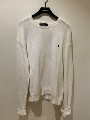 Vit stickad tröja från Polo Ralph Lauren - Klassisk vit stickad tröja från Polo Ralph Lauren med rund halsringning och diskret logga broderad på bröstet. Tröjan har ribbade muddar vid ärmslut och nederkant, och är tillverkad i mjukt material som känns skönt mot huden. Har en liten fläck som borde gå bort i tvätten. 