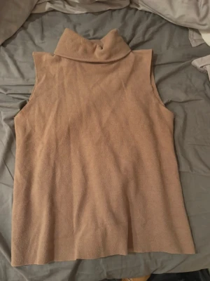 Beige ärmlös polotröja från Zara - Säljer en beige/ brun ärmlös polotröja från Zara. Tröjan har en hög krage och är tillverkad i ett mjukt, stickat material. Perfekt att styla lager på lager eller bära som den är för en clean look.