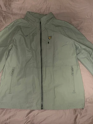 Grön vindjacka från Lyle & Scott - Snygg grön vindjacka från Lyle & Scott med dragkedja framtill, två sidofickor med dragkedja och justerbar huva. Jackan har det klassiska Lyle & Scott-logotypen på bröstet och är tillverkad i lätt polyester, perfekt för blåsiga dagar.