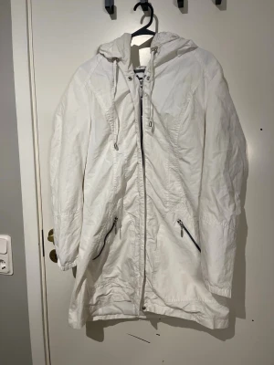 Vit parkas med huva och dragkedja - Vit parkajacka med huva och snörning, dragkedja framtill och två fickor med dragkedja. Jackan har lång ärm och en rak, något längre passform. Perfekt för kyligare dagar och enkel att matcha med olika outfits.