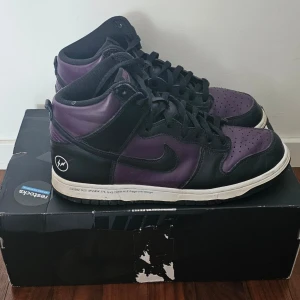 Nike Dunk High svart/lila sneakers - Nike Dunk High sneakers i svart och lila med klassisk snörning och vit sula. Skorna har en hög siluett och är tillverkade i skinn med detaljer i syntet. Perfekta för dig som vill sticka ut med en unik färgkombo och ikonisk design.