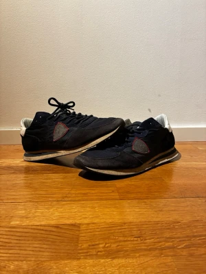 Philippe model - Snygga marinblåa sneakers med vita detaljer och röd kant runt loggan på sidan. Skorna har snörning och platt sula, samt en klassisk låg siluett. Materialet är en mix av mocka och syntet, vilket ger en sportig och stilren look.