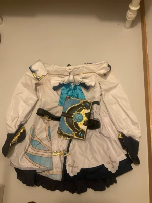 Barbara cosplaydräkt med peruk - Cosplaydräkt inspirerad av Barbara från Genshin Impact. Inkluderar vit och blå klänning med guldiga detaljer, bälte med bokaccessoar samt blond vågig peruk och huvudbonad. Perfekt för cosplay och maskerad.og pris 1700