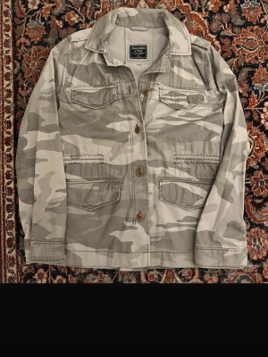 Camouflage overshirt från Abercrombie - Snygg overshirt från Abercrombie & Fitch i beige och grått camouflage-mönster. Jackan har fyra fickor framtill, knäppning med träknappar och klassisk krage. Perfekt lager-på-lager-plagg för en avslappnad streetstil.