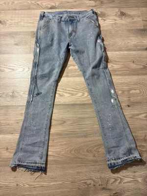 Sumwon Flared Jeans - Säljer ett par ljusblå/grå Sumwon flared jeans med slitna detaljer och råa benslut. Jeansen har vita färgstänk och dekorativa snören längs benen för en edgy look. Perfekta för dig som gillar streetstyle och vill sticka ut.