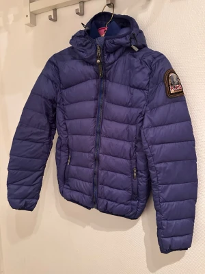 Blå dunjacka från Parajumpers - Snygg blå dunjacka från Parajumpers med huva och dragkedja framtill. Jackan har ett broderat märke på ena ärmen och två fickor med dragkedja. Perfekt för kalla dagar och riktigt skön att bära.