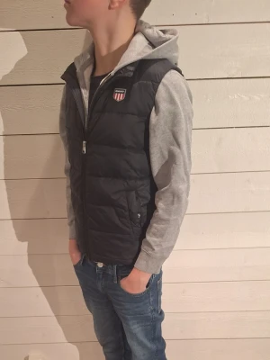 Svart dunväst från Gant - Säljer en svart dunväst från Gant med quiltad design och dragkedja framtill. Västen har en klassisk passform och en liten logotyp på bröstet. Perfekt att slänga över en hoodie för en chill look. Modelen är 162cm
