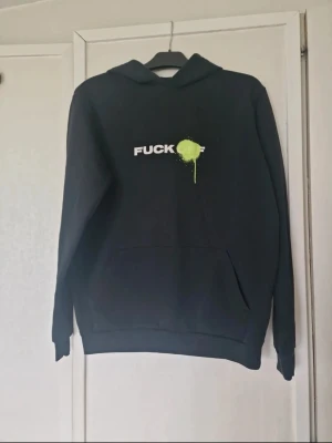 Svart hoodie med tryck och huva - Svart hoodie med vit text "fuck off" och neongrön färgdetalj framtill. Klassisk modell med huva och magficka. Den är nopprig och saknar snören till huvan.