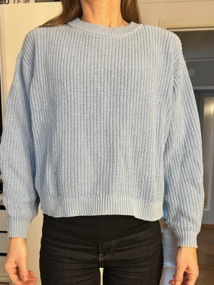 Ljusblå stickad tröja i bomull - Säljer en ljusblå stickad tröja med rund hals och avslappnad passform. Tröjan har ribbade muddar vid ärmslut och nederkant samt en grovstickad struktur. 