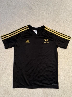 Svart Adidas Salah tröja med guld, nr 10. - Snygg svart sporttröja från Adidas med guldiga detaljer och Salah-tryck på bröstet. Tröjan har tre guldiga ränder på axlarna, Adidas-logga och Salahs namn samt nummer 10 i guld på ryggen. Tillverkad i lätt och ventilerande material, perfekt för träning eller match. Tvättrådet bortklippt annars mycket fint skick.