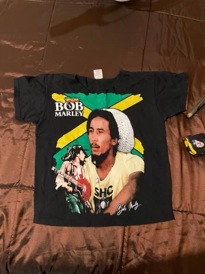 Svart Bob Marley t-shirt med tryck - Cool svart t-shirt med stort färgglatt Bob Marley-tryck på framsidan och texten 'BOB MARLEY' i gult på baksidan. Klassisk passform och korta ärmar. Perfekt för dig som gillar musik och vill sticka ut med en ikonisk look..barn storlek (8-9år)