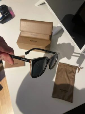Burberry svarta solglasögon - Snygga svarta solglasögon från Burberry med fyrkantig form och klassiska ränder i vitt, rött och beige på skalmarna. Märkeslogga på sidan. Kommer med originalfodral och tygpåse. Perfekta för dig som vill ha en stilren och trendig accessoar.