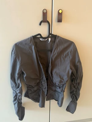 Brunb figur kavaj från Weekday - Snygg mörkgrå blus från Weekday med långa ärmar och rynkade detaljer i midjan. Blusen har djup v-ringning och knäppning framtill med små knappar. Tillverkad i ett tunt, lätt bomullstyg som ger en avslappnad men trendig look.