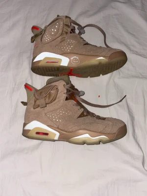 Nike Air Jordan 6 Travis Scott sneakers - Unika Nike Air Jordan 6 Travis Scott sneakers i beige mocka med vita och röda detaljer. Skorna har snörning, ficka på sidan och en platt sula. Kommer med matchande brun tygpåse med röd text. Perfekt för dig som vill sticka ut.