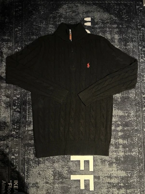 Svart kabelstickad tröja Polo Ralph Lauren - Kabelstickad svart tröja från Polo Ralph Lauren med halv dragkedja och röd broderad logga på bröstet. Tröjan har hög krage, långa ärmar och ribbade muddar. Perfekt för dig som gillar klassisk och stilren design.