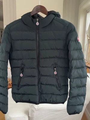 Svart pufferjacka med huva från Colmar - Snygg svart pufferjacka från Colmar  med huva och quiltad design. Jackan har dragkedja framtill och på fickorna, samt logga på ärmen. Tillverkad i syntetmaterial och perfekt för kyliga dagar.