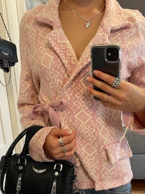 Rosa omlottkofta - Superfin stickad kofta i ljusrosa och vitt från Lely Wood med snyggt geometriskt mönster, krage och knytband. Finns ett litet hål (sista bilden), men är på insidan av armen så syns inte vid användning, men går annars lätt att sy! Är justerbar i midjan som ger en perfekt passform, passar storlek S/M.