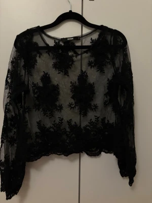 Svart transparent spetsblus Bik Bok - Snygg svart transparent blus från Bik Bok med långa ärmar och detaljerat blommigt spetsmönster. Blusen har rund halsringning och är perfekt för att skapa en trendig lager-på-lager-look. Materialet är tunn mesh med broderade spetsdetaljer.