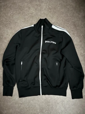 Palm Angels svart track jacket - Säljer en svart track jacket från Palm Angels med vita ränder längs ärmarna och vit dragkedja framtill. Tröjan har hög krage, två fickor med dragkedja och logga på bröstet. Tillverkad i polyester för en sportig vibe.