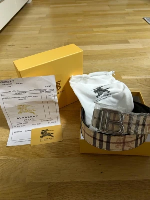 Burberry bälte med klassiskt rutmönster - Snyggt Burberry-bälte i beige, svart och rött med det ikoniska rutmönstret och en stor silverfärgad B-spänne framtill. Bältet är gjort i läder och canvas, och levereras med originalpåse och box. Perfekt accessoar för att lyfta din outfit.