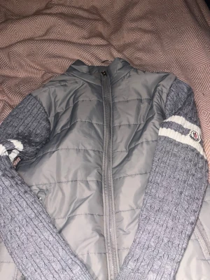 Grå cardigan jacka från Moncler - Snygg grå cardigan jacka från Moncler med quiltad kropp i polyester och stickade ärmar. Ärmarna har vita ränder och Moncler-logga på vänster arm. Dragkedja framtill och hög krage för extra stilpoäng.