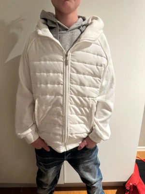 Vit vadderad vindjacka med huva - Snygg vit vindjacka med quiltad framsida och baksida, dragkedja och huva. Jackan har en sportig look med raka linjer och två fickor framtill. Perfekt för dig som vill ha en stilren och enkel jacka till vardags.