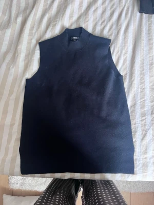 Marinblå stickad väst från Uniqlo - Säljer en marinblå stickad väst från Uniqlo med hög krage och ärmlös design. Västen har en rak passform och är gjord i mjukt material som känns skönt mot huden. Perfekt att styla över en skjorta eller t-shirt för en clean look.
