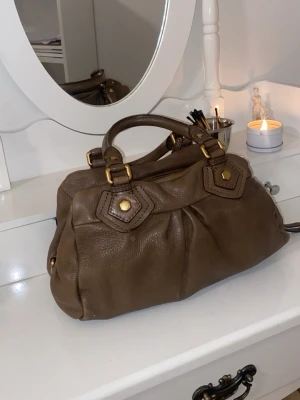 Brun Marc Jacobs handväska - Säljer en snygg brun handväska från Marc Jacobs i mjukt skinn. Väskan har guldfärgade metalldetaljer, dubbla handtag och en dekorativ metallbricka framtill. Fint skick och super gullig. 
