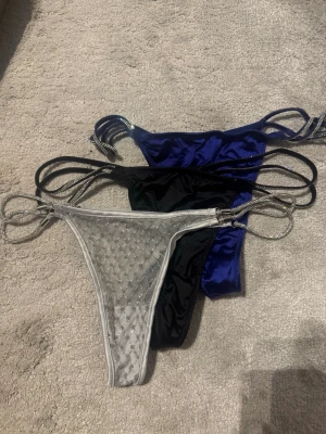 3-pack stringtrosor med glitterband - Säljer ett 3-pack stringtrosor i silver, svart och blått. Alla har tunna, glittriga band i sidorna och en sexig, minimalistisk design. Den silvriga trosan är i mesh med diskret mönster, medan de svarta och blå är i glansigt tyg. Perfekta för dig som gillar trendiga detaljer.