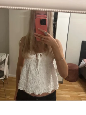 Vit volangtop med knyt fram - Säljer en söt vit topp med volangkant och knyt fram. Toppen har smala axelband och är gjord i ett strukturerat, stretchigt material som ger en luftig känsla. Perfekt för varma dagar och enkel att styla med jeans eller kjol.