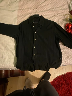 Svart skjorta med knappar - Säljer en svart skjorta med klassisk krage och vita knappar framtill. Skjortan har lång ärm och en loose passform, perfekt för en avslappnad stil. Tillverkad i ett mjukt tyg som känns skönt mot huden.